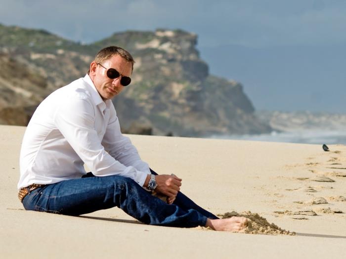 بنطلون جينز غير رسمي ضيق ، وقميص أبيض ، يرتديه دانيال كريج ، مع نظارة شمسية أفياتور ، جالسًا حافي القدمين ، على شاطئ رملي