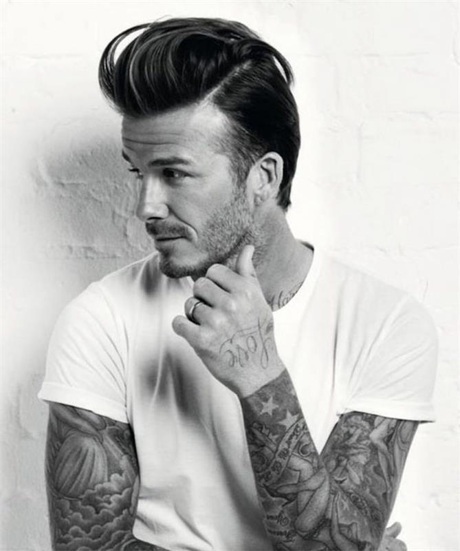 David Beckham v bielom tričku, s tetovaním na ruke a s strniskovou bradou, vlasmi s podrezanými vlasmi a pompadourom