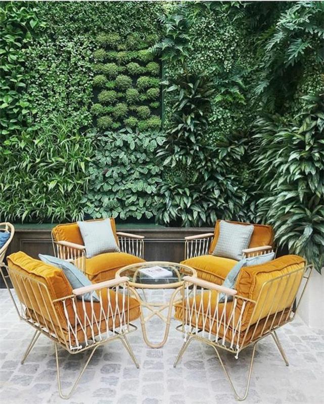 vonkajšie usporiadanie domu, vonkajšia dekorácia steny, zelená stena, ktorá kombinuje niekoľko druhov rastlín, záhradný nábytok so štyrmi kreslami s bielymi kovovými rámami s oranžovými zamatovými vankúšmi, malý okrúhly stôl so sklenenou doskou