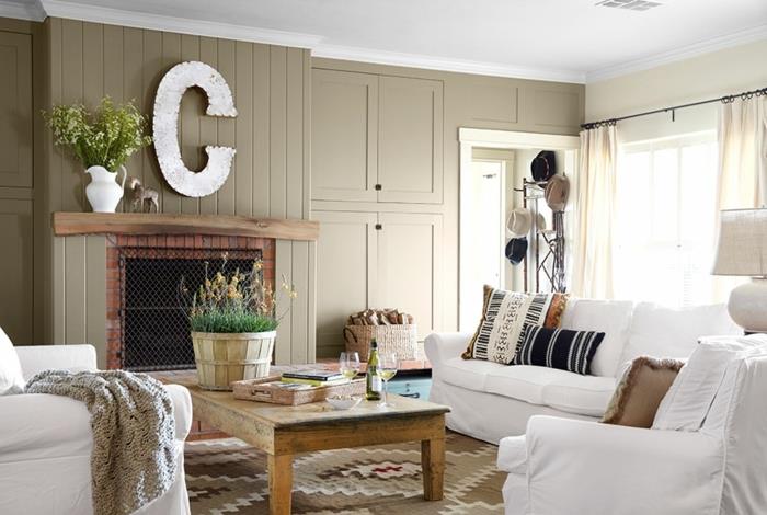 soffbord i rått trä, vit soffa och fåtöljer, dekorativt väggbrev, öppen spis, mönstrad brun matta, mångfärgade kuddar, rustik samtida inredning