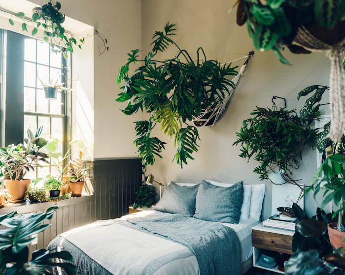 výzdoba spálne v tropickom štýle Tumblr so zelenou vegetáciou, zelené rastliny na okne, rebrod a kvetináče visiace na stene s macrame, sivou a bielou posteľnou bielizňou