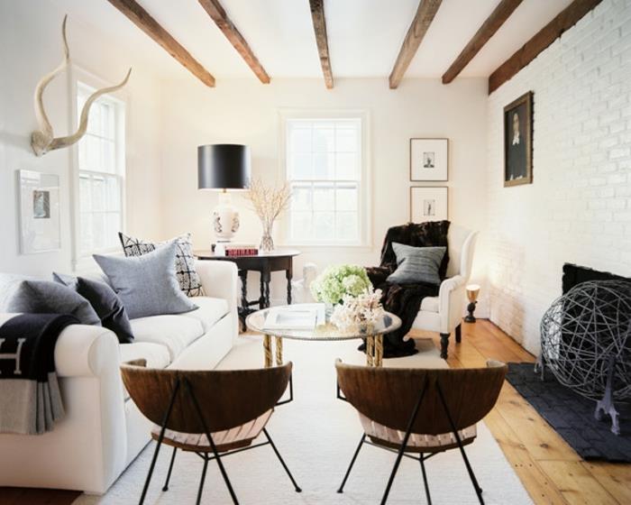 rustik chic landsbygdsdeo, trästolar, vit soffa, grå kuddar, runt glas- och metallbord, vit matta, öppen spis, vit tegelvägg, exponerad balk, hjorttorn väggdekoration