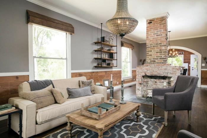 Rustik öppen spis i tegelsten, grå matta med geometriska mönster, beige soffa, grå fåtölj, soffbord i trä, designerlampa, grå vägg, chic country -inredning