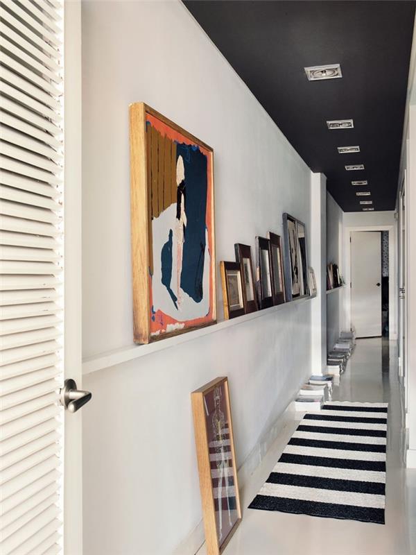 lång och smal korridordekoration i svart och vitt med en hylla med konstnärliga ramar och ett matt svartmålat tak för en fin grafisk effekt