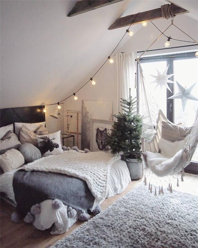 dekor mix rustikálny a moderný škandinávsky dekor spálňa pod strechou odhalené trámy hojdačka závesná posteľ zdobená sivým a bielym ľanovým vencom zo žiaroviek