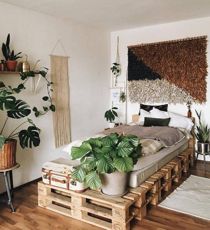 bohémska elegantná výzdoba spálne s vlastnou paletovou posteľou, sivým matracom, koncom postele v črepníkovej rastline a vintage kufrom, monstera deliciosa na servírovacom stole, macrame a tkaním na stenu