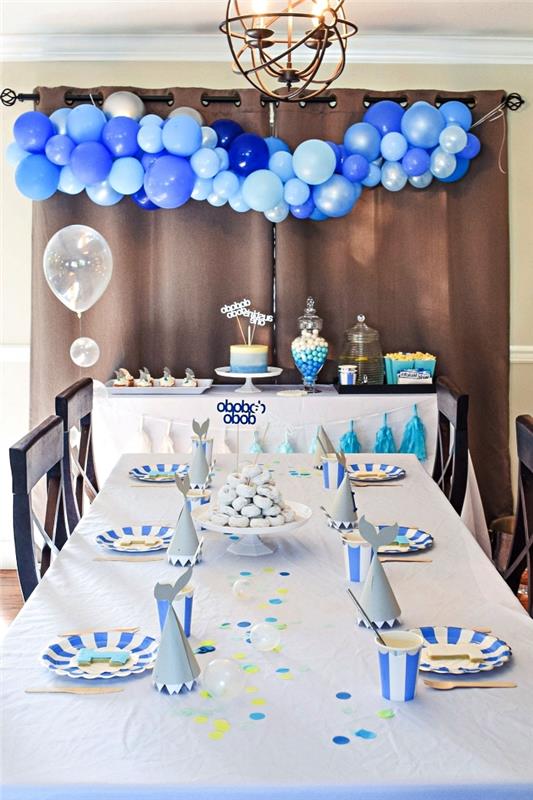 1 års jubileumsborddekoration i blått och vitt, konfetti mittpunkt och bordduk med vita och blå ränder, födelsedagsbuffé dekorerad med en ballongbåge i blå nyanser
