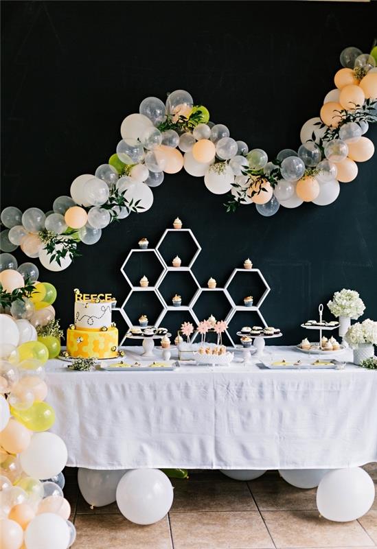 bi -födelsedagstema i svart, gult och vitt för att fira din bebis första år, en födelsedagsborddekoration med ballonger och konstgjorda blad, födelsedagsbuffé med godislådor och muffinsdisplay i form av en bikakebi dekorerad med en ballongbåge på en svart bakgrund
