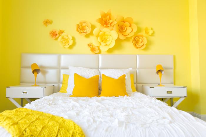 žltá farba spálne pre dospelých, bielo -žltá posteľná bielizeň, papierová dekorácia na stenu, biely nočný stolík