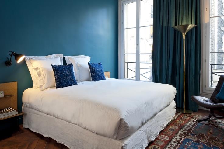 Nápad na výzdobu spálne pre dospelých, tmavomodrá akcentovaná stena, pávový modrý záves, biela posteľná bielizeň s modrými ozdobnými vankúšmi, orientálny koberec, hnedé parkety