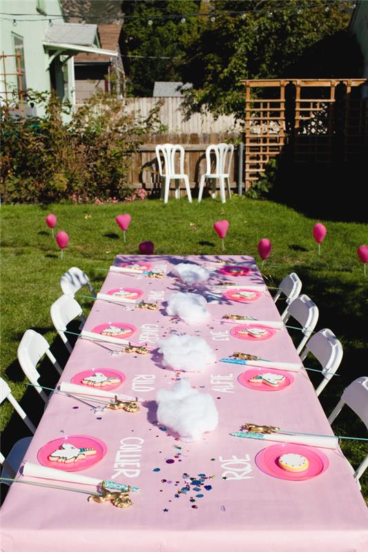 dekorativa idéer för en utomhusfödelsedagsfest, vacker bordsdekoration med en personlig rosa bordsduk med namnen på gästerna skrivna på den och ett enhörningstillbehör nära varje tallrik