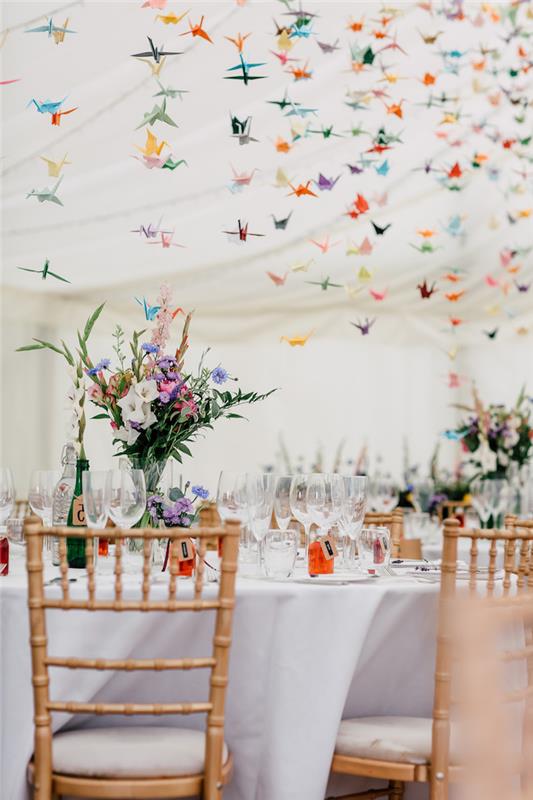 origami farebné papierové žeriavy visiace zo stropu a kyticová výzdoba farebných vidieckych kvetov na bielych obrusoch, farebná výzdoba svadobnej miestnosti