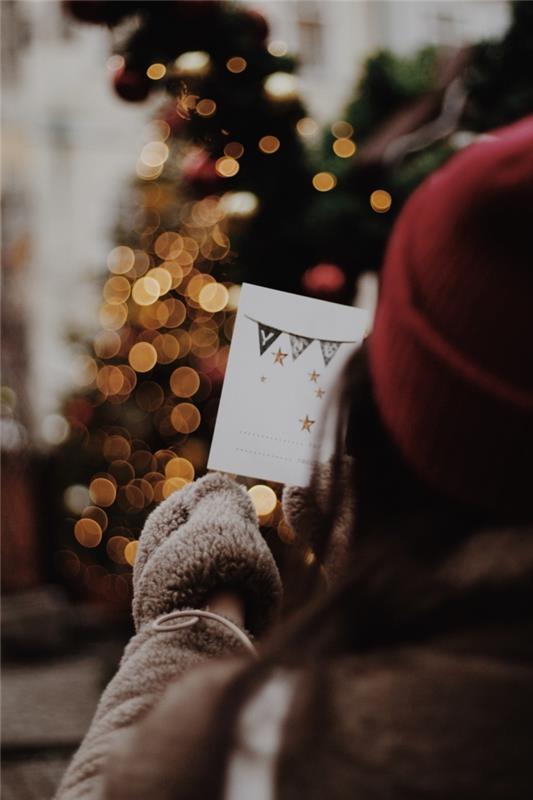veselý vianočný obraz na tapetu pre smartphone, dievča oblečené v umelom kožuchu a červenej šiltovke s vianočnou pohľadnicou v rukách pred stromom