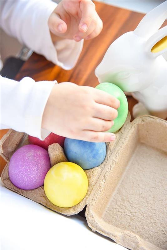يد صغيرة تمسك قطعة من السلايم الخضراء على شكل بيضة ، فوق كرتونة بيض ، تحتوي على بيض أحمر وأزرق وأرجواني وأصفر ، كيفية صنع السلايم بالغراء