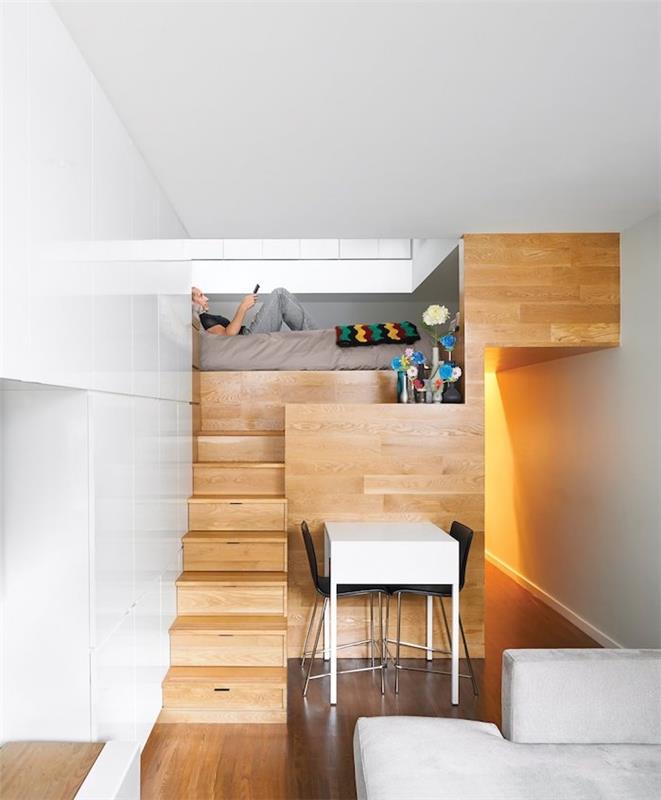 minimalistisk design i en liten trästudio med säng ovanför ett kök, vitt bord och svarta stolar, ljusgrå soffa