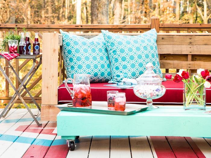 príklad záhradného nábytku v palete s drevenou sedačkou s červeným vankúšom na sedenie, modrými ozdobnými vankúšmi a konferenčným stolíkom v palete premaľovaným na modrú farbu s kolieskami, deco terasa