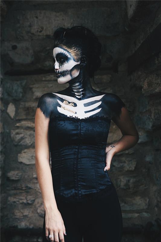 žena s kostlivým halloweenskym make -upom oblečená v čiernej tvári s polovičnou lebkou