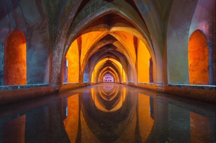 de underjordiska gångarna i en gotisk kyrka, grå bågar upplysta av ett gult ljus, mystiskt utrymme, vattenkanal som reflekterar bågarna