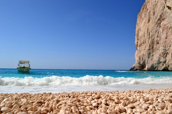 båt som närmar sig stranden, blått vatten, paradisiskt landskap, leraberg som stiger på stranden, små ovala vita stenar på stranden