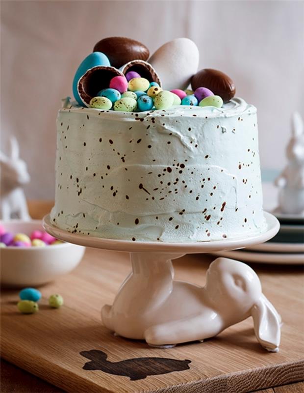 fläckig påskboxtårta med pastellgrön färgad smörkrämfrosting dekorerad med chokladägg, placerad på ett sött kaninkakstativ