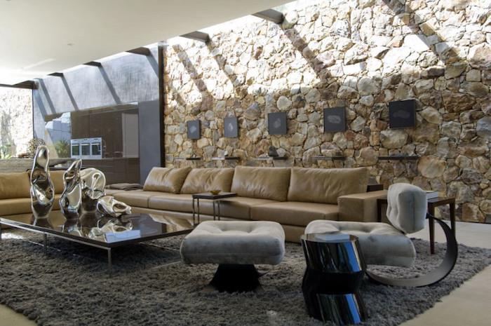 veľká dizajnérska obývačka s vnútornou kamennou stenou a falošným stropom s odhalenými trámami a veľkou pohovkou z béžovej kože