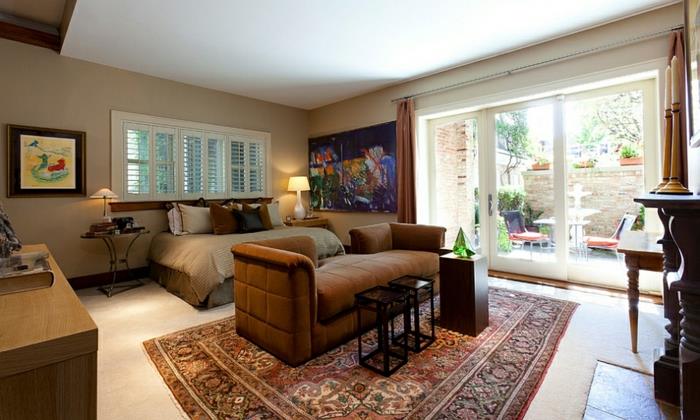 غرفة نوم مع طلاء محايد ، رمادي داكن وأبيض ، سجادة فارسية ، أريكة بنية ، طاولات صغيرة ؛ جدار زجاجي