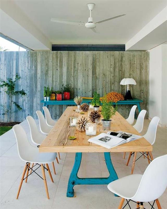 dekorácia exteriérovej steny, ozdobenie vonkajšej steny, leštená sivá drevená krytina, vintage stôl natretý tyrkysovou modrou nalepený na stenu, záhradný nábytok s veľkým obdĺžnikovým stolom so žltou drevenou doskou a kovové nohy natreté tyrkysovo modrou farbou, osem stoličiek z bieleho plastu s operadlami a tenké nohy z ľahkého dreva