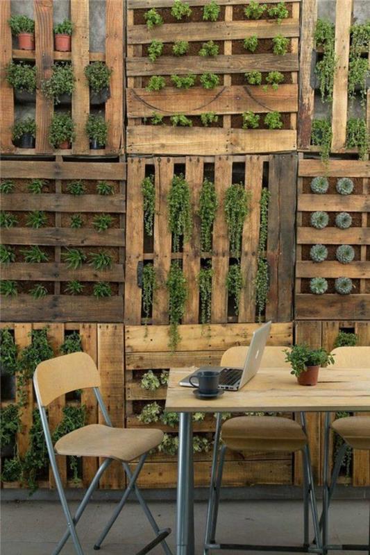 dekoratívna vonkajšia stena z dreva z hnedých paliet s vegetáciou, ktorá vychádza cez otvory v paletách, zelená stena na terasu alebo do záhrady, veľký štvorcový stôl s doskou zo svetlého dreva, tri skladacie stoličky z PVC dreva a nohy v hliníkových trubkách