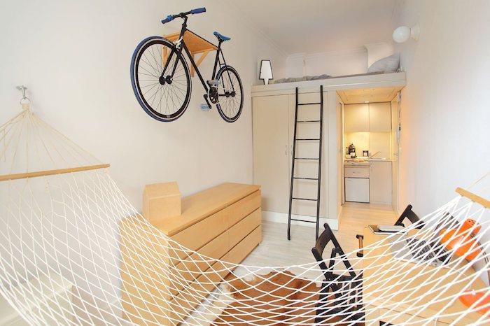 vita väggar och minimalistisk design i en 20 m2 studio, studiokök och garderob med säng ovanför, hängmatta inomhus, skrivbord och byrå i trä