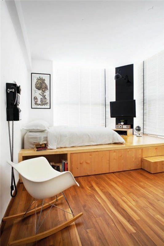 dekorácia spálne pre dospelých s posteľou na platforme a bielym plastovým hojdacím kreslom, časť steny natretá na čierno, na ohraničenie televízneho priestoru, podlahová krytina vo farbe whisky, masívne parkety