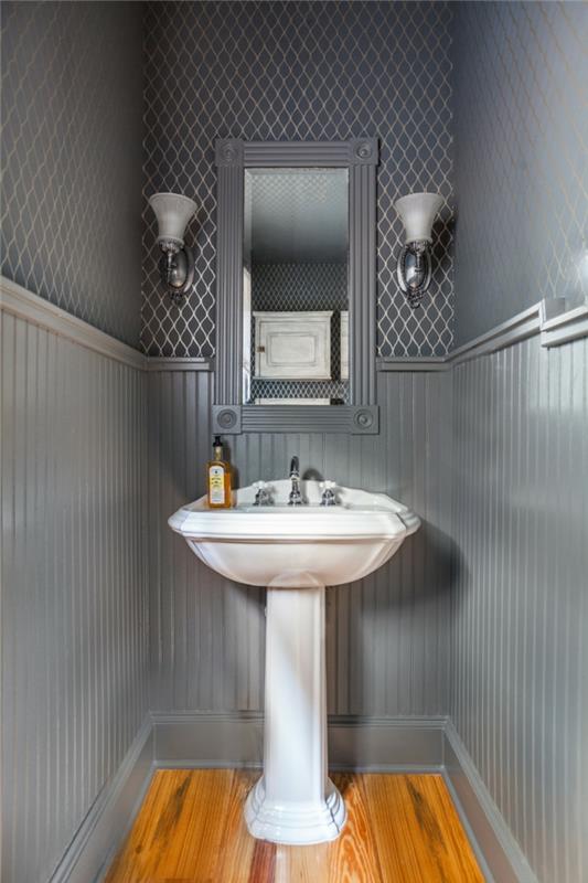 WC v šedej farbe, biele podstavce, dve nástenné svietniky a zarámované zrkadlo, geometrická tapeta na stenu, drevená podlaha