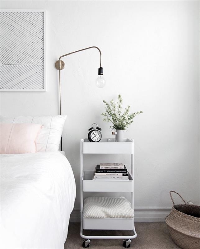 Nápad na výzdobu škandinávskej spálne v bielej farbe s ružovým vankúšom a bielym nočným stolíkom na kolieskach s policami, sivým kobercom