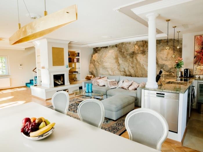 idé om att ordna ett litet kök med vit centralö och bänkskiva i marmor, som vetter mot ett vardagsrum med stenstenad accentvägg, grå hörnsoffa, soffbord och orientalisk matta, original hängande ljus, matbord och stolar