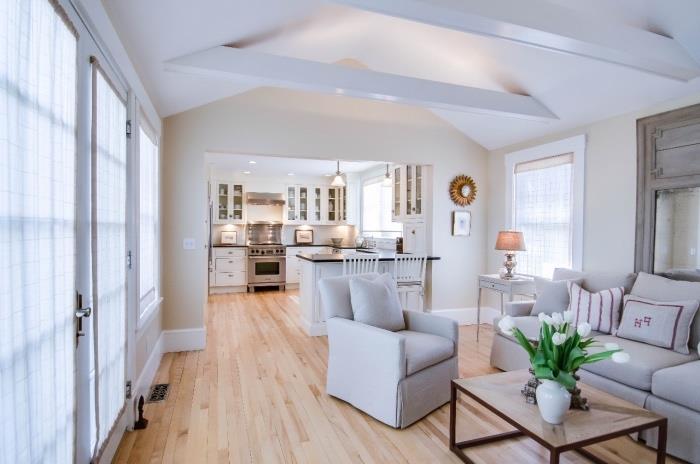 möblera ett litet klassiskt vitt kök som vetter mot ett vardagsrum med vita synliga balkar, grå soffa och fåtölj och soffbord i trä, ljus parkett