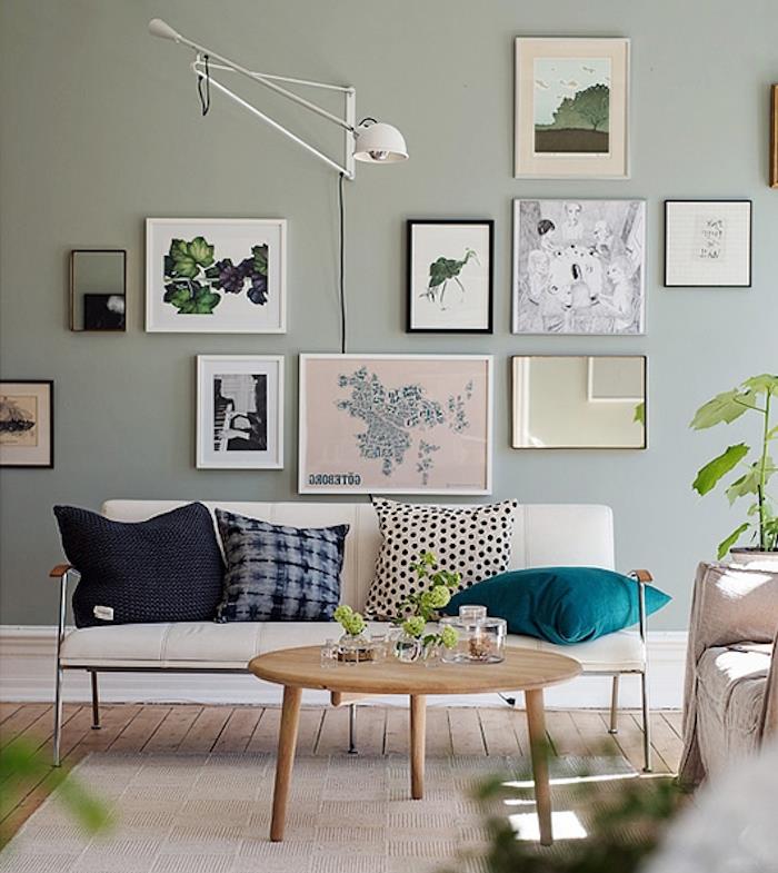 stena premaľovaná na pastelovo zelenú farbu, celadonový odtieň v dizajnérskej obývačke so slonovinovou sedačkou, ozdobená modrými, sivými a čiernymi vankúšmi, drevený konferenčný stolík, svetlo šedý koberec, stena z ozdobných rámov, zelené rastliny