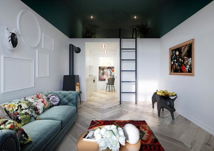 mörkgrönt tak och vita väggar, litet kök i vitt, stor påfågelblå soffa dekorerad med blommiga kuddar, färgglad matta, väggar dekorerade med konstmålningar
