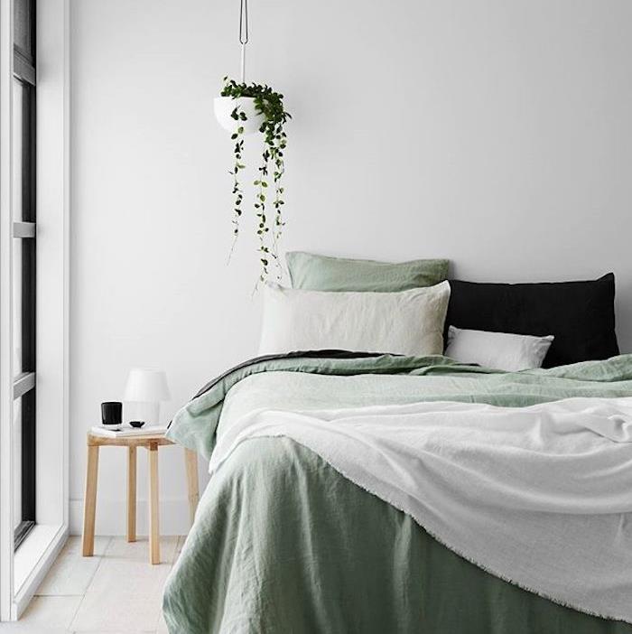 dekorácia spálne pre dospelých, biela stena, biela a zelená posteľná bielizeň, čierny vankúš, svetlé parkety, drevený nočný stolík, závesná zelená rastlina
