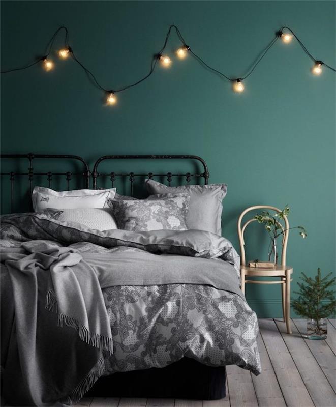 výzdoba hlavnej spálne s tmavozelenou farbou na stenu a dekorom svetlého vence, drevenou podlahou, bielou a sivou posteľnou bielizňou na čiernej kovovej posteli