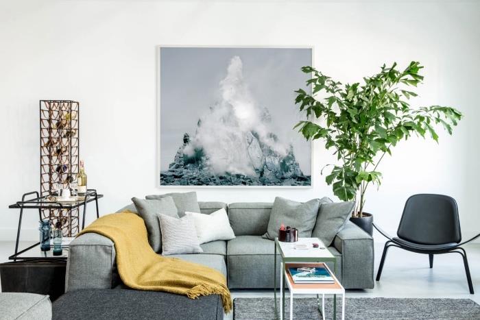 šedá pohovka a koberec, okrová žltá deka, biela podlahová a stenová krytina, zelená rastlina, dekoratívna rámová maľba na stenu