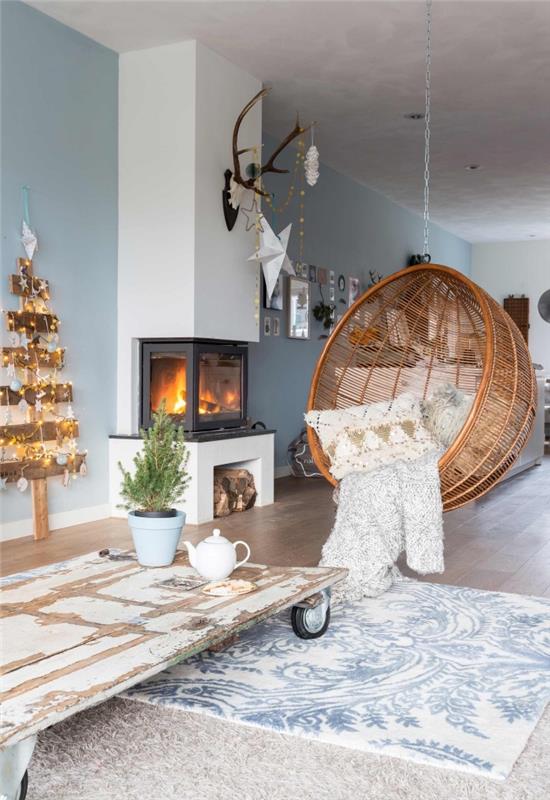 kokande skandinavisk vardagsrumsinredning i vitt och blågrått som inbjuder dig att koppla av tack vare sin hängande rottingstol och juldekorationen och dess julgran i trä dekorerad med små ljusa kransar