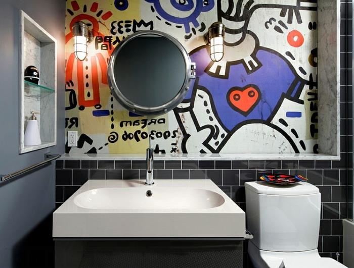 čierne toaletné dlaždice metra, originálna nástenná maľba s farebnými vzormi, okrúhle zrkadlo, biele umývadlo na dosku, sivá stena, dve priemyselné svietniky