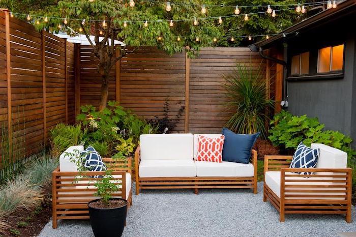 terrass i terrass med soffa och fåtöljer i trä, grus, växtgräns, sittdynor, vita ryggstöd, träd och ljusa kransar