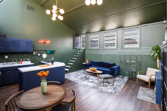 olivgröna väggar, kök, centralö och mörkblå soffa, mörkbrun parkett, runt träbord omgivet av antika stolar, grå och vit matta