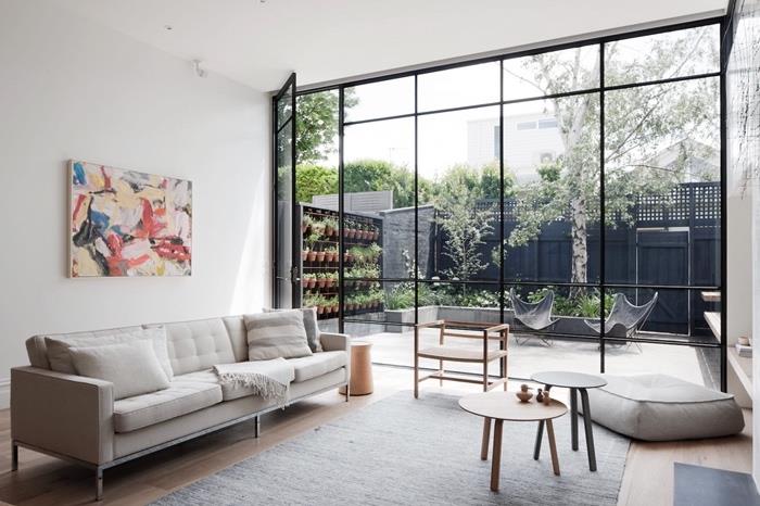 Obývačka inšpirovaná Škandináviou, sivý koberec, biela pohovka, drevené hniezdne stolíky, sivý pohovka a presklená strecha s výhľadom do záhrady