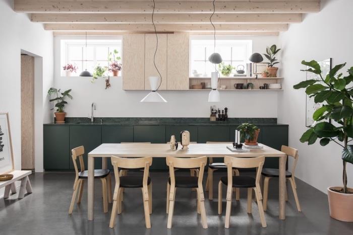 Škandinávsky nábytok, masívny drevený stôl, drevené a čierne kožené stoličky, tmavozelená kuchynská časť a odhalené trámy, dekorácia zelených rastlín