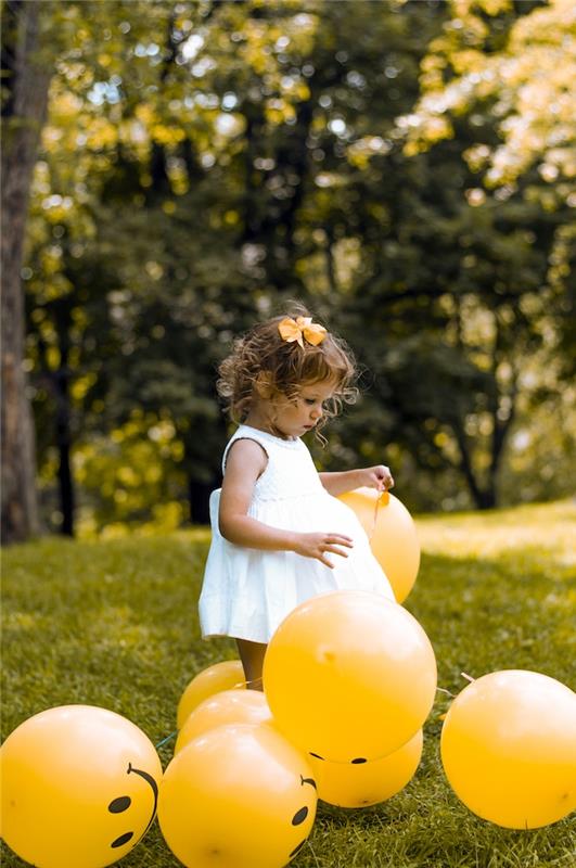Prvá narodeninová párty so žltými balónikmi, záhradné dievča hrajúce sa s balónikmi, dievčatko v bielych šatách, účes s kučeravými vlasmi, roztomilý žltý motýľ
