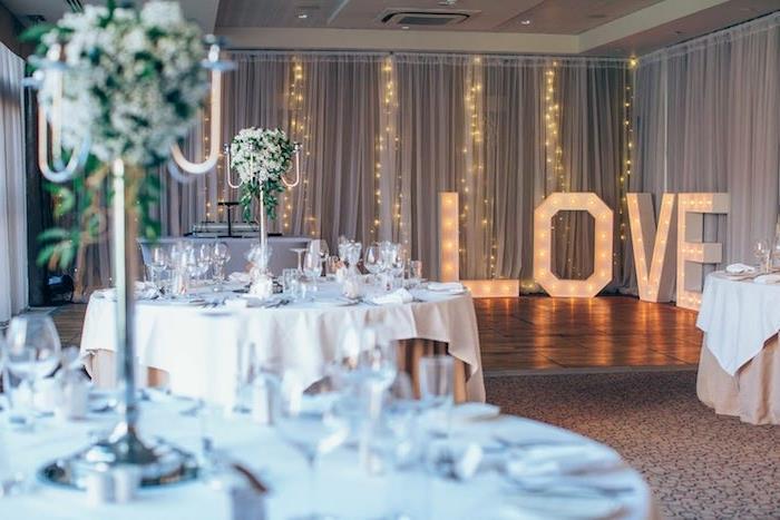 romantická svadobná téma so svetlou girlandovou výzdobou, svietiacimi písmenami, kyticami bielych kvetov na stoloch s bielym a béžovým obrusom