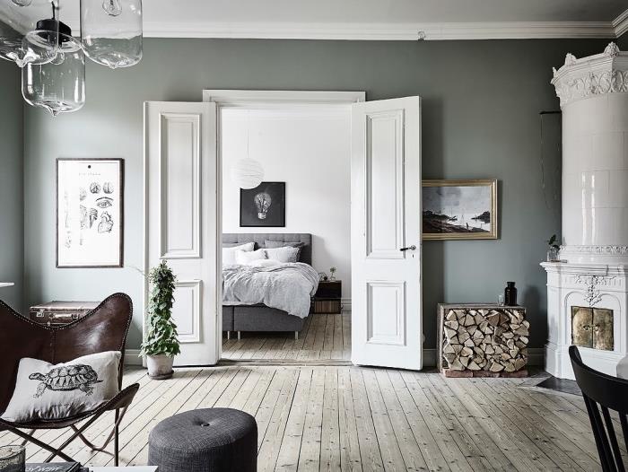 spálňa otvorená do obývačky, svetlo sivé parkety, sivá posteľ s bielou posteľnou bielizňou, kožená stolička, biely krb, rustikálny interiér