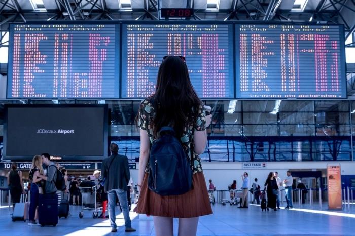ung flicka framför flygplatsstyrelsen, stort väntrum, flygplats, ryggsäck, terrakottafärgad kjol