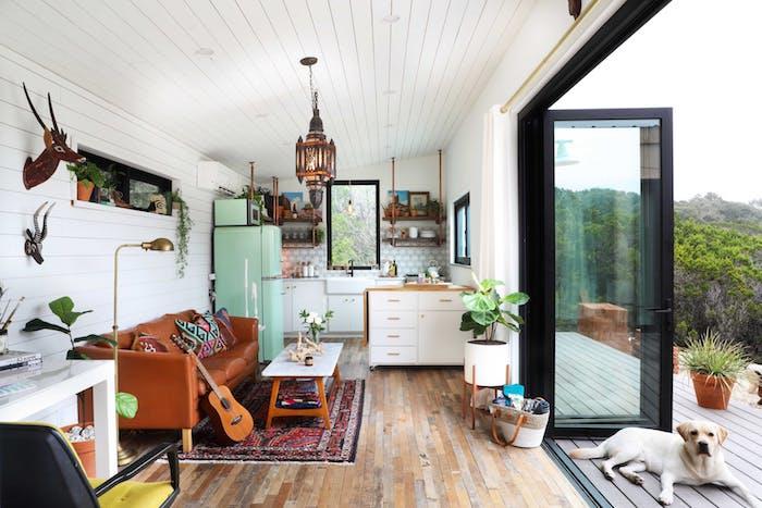bohemisk chic stil deco i ett kök öppet för vardagsrum med brun skinnsoffa, orientalisk matta, grönt kylskåp, vitt kök med liten ö, gröna växter inomhus